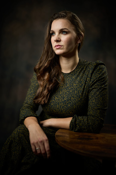 Woman in olive-green knit sweater posed against dark background with hand resting on wooden surface.