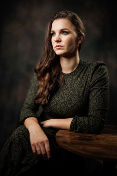 Woman in dark green knit sweater with long brown hair, posed against dark textured background.