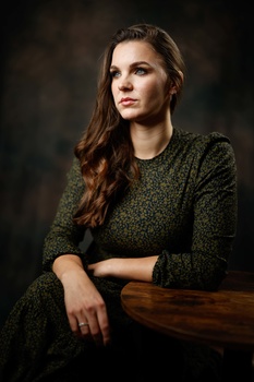Woman in olive knit sweater with long brown hair, posed against dark background with hand resting on wooden surface.