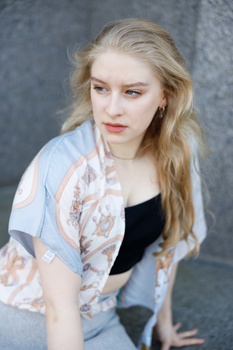 Young woman with long blonde hair wearing a patterned cardigan over black top, photographed against gray backdrop.