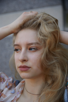 Young woman with wavy blonde hair looking downward, hand raised to head, against blurred gray background.