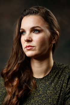 Woman with long brown hair wearing a textured olive-green shirt, photographed against a dark blurred background.
