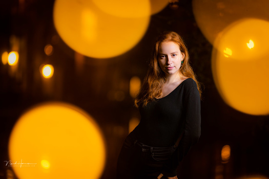 Woman with reddish-blonde hair wearing black clothing, positioned in center frame with blurred golden orb lights in background.