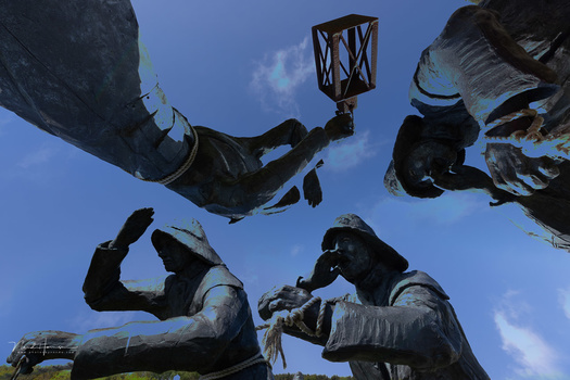 Four divers in wetsuits and gear positioned around a large underwater structure, viewed from below against a blue sky.