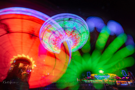 Nighttime long-exposure photograph of illuminated carnival rides with vibrant red, green, and blue neon lights creating motion blur.