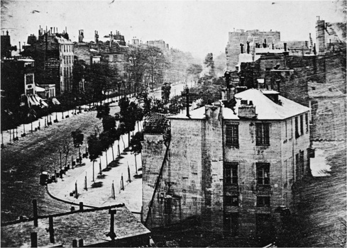 Historical black and white cityscape with tree-lined street and multi-story buildings receding into distance.