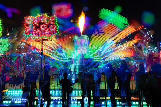 Silhouetted crowd standing before a vibrant neon-lit carnival or fair scene with colorful light displays and signs.