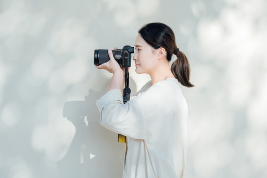 Woman in white clothing holding a compact camera up to her face against a bright, overexposed background.