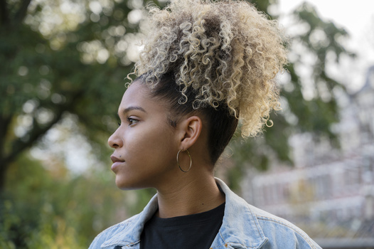 Woman with bleached blonde curly hair in a top knot, wearing a denim jacket over a black shirt, photographed outdoors in profile.