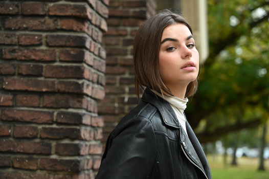 Young woman with long dark hair wearing a black leather jacket, looking over her shoulder against a brick wall.