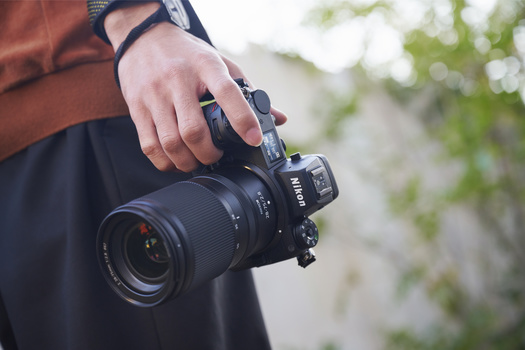 Photographer holding a Nikon DSLR camera with telephoto lens in both hands outdoors.