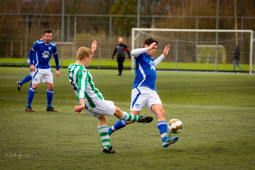 Young soccer player in green and white striped kit kicking ball while defender in blue jersey extends arms.