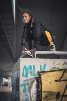 Person in dark jacket posing on graffitied concrete structure under bridge overpass.