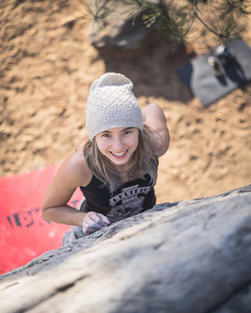 Young woman in a beanie climbing a sandy rock face, smiling at the camera from above.