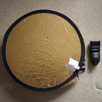 Gold and silver 5-in-1 reflector with a camera flash positioned beside it on a gray surface.