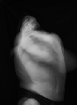 Black and white artistic photograph of a muscular male torso and shoulder, captured with soft directional lighting against a dark background.