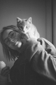 Person wearing glasses holding a cat on their shoulder, photographed in black and white.