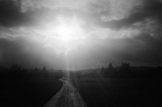 A straight road stretches toward a bright light breaking through dark, heavy clouds in a dramatic black and white landscape.