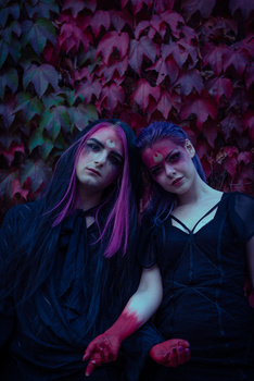 Two people posing against a vibrant magenta ivy-covered wall in moody purple-tinted lighting.