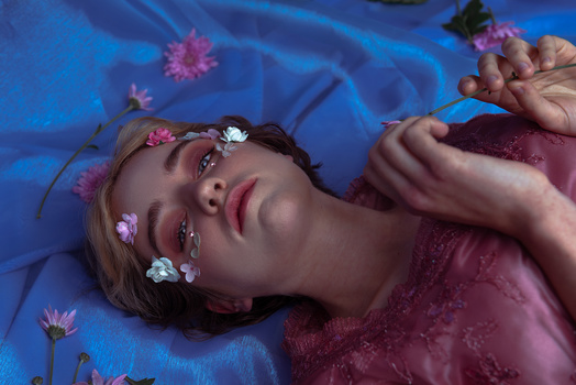 Woman lying on blue fabric with pink flowers and glitter makeup adorning her face and eyes.