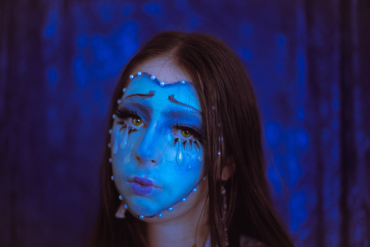 Portrait of a woman with blue face paint and dramatic eye makeup against a dark blue background.