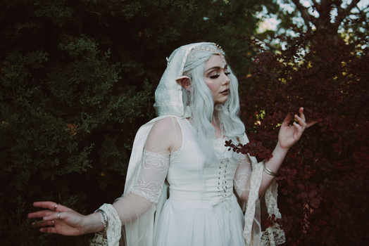 Woman with long white hair in white medieval-inspired dress surrounded by dark green foliage.