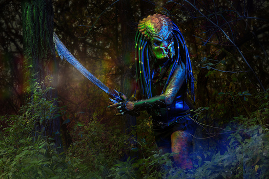 Person in neon body paint and dreadlocks holding an illuminated sword in a forest setting.