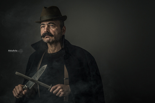 Man in black fedora and jacket holding two large knives against dark background.