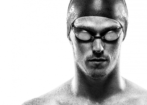 Shirtless swimmer wearing goggles and swim cap, photographed against white background with water droplets on skin.