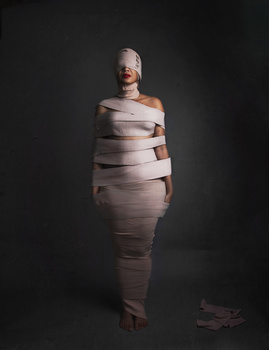 Person wrapped in white gauze bandages against a dark background, posed standing upright.
