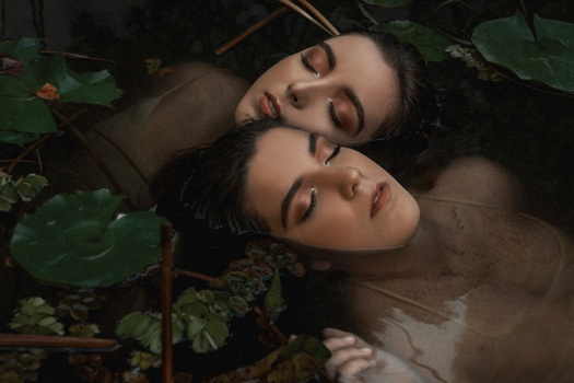 Two women with closed eyes resting among foliage and water in soft, warm lighting.