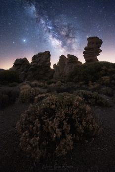 Milky Way galaxy arching over desert rock formations and shrubland at night.