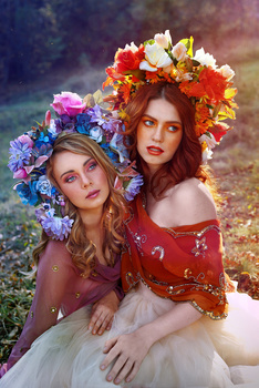 Two women wearing elaborate floral crowns pose together outdoors with soft, warm lighting.