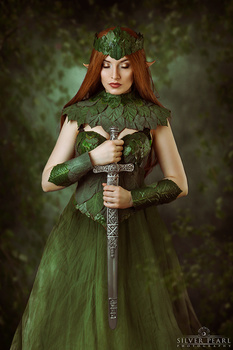 Woman with long red hair wearing a green tulle gown and crown, holding a decorative sword with ornate hilt.