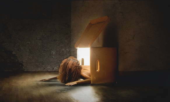 Person with curly hair emerging from an open wooden box emitting bright light against a dark concrete interior.