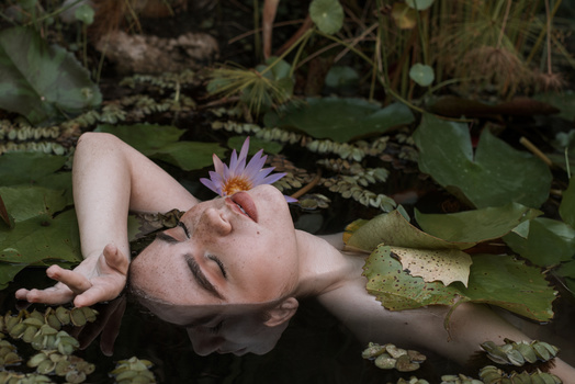 Person's face submerged in still water surrounded by lily pads and a purple water lily flower.