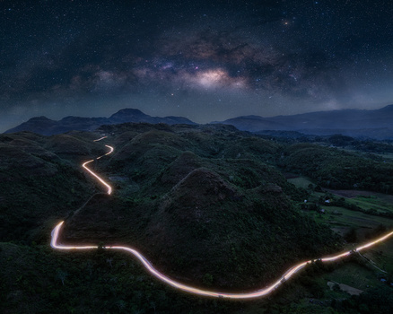 Winding road with car light trails beneath the Milky Way and mountains at night.
