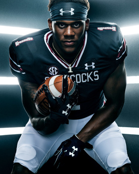 Football player in dark uniform holding football in athletic pose under dramatic lighting.