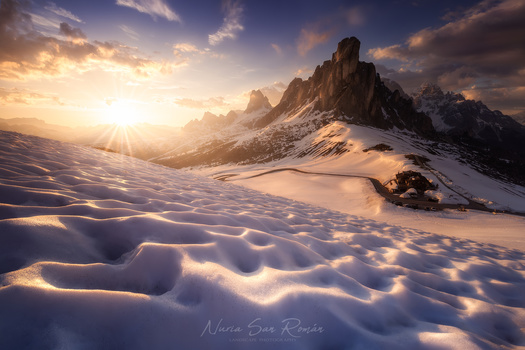 Snow-covered alpine landscape with jagged mountain peaks glowing in golden sunrise light.