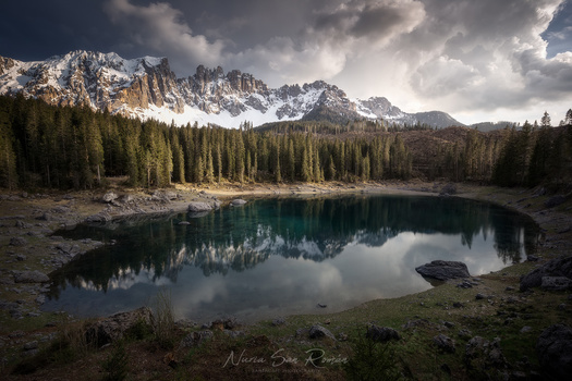 Alpine lake with mirror-like reflection surrounded by coniferous forest and snow-capped mountain peaks under dramatic cloudy sky.