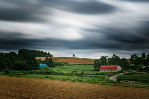 Rural farmland with red barn, rolling green fields, and dramatic overcast sky.