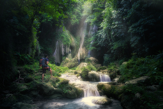 Hiker with backpack walking toward a cascading stream through a lush, moss-covered forest canyon.