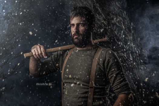 Bearded man holding an axe against a stone wall in falling snow.