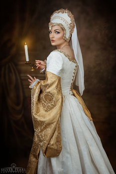 Woman in period costume holding a candle, wearing white linen dress with gold embroidered fabric drape and ornate headdress.