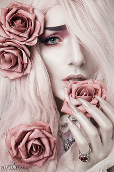 Portrait of a woman with platinum blonde hair adorned with dusty rose flowers, pale complexion, and decorative rings.