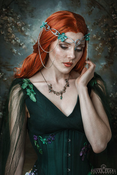 Woman with long copper-red hair wearing an elaborate green dress and ornate headpiece adorned with chains and crystals.