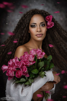 Woman with long curly dark hair holding pink roses, wearing white garment against dark background with floating petals.