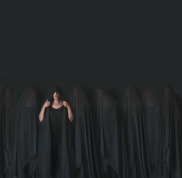 Woman in black top stands centered among draped dark fabrics, gazing downward with hands near shoulders.