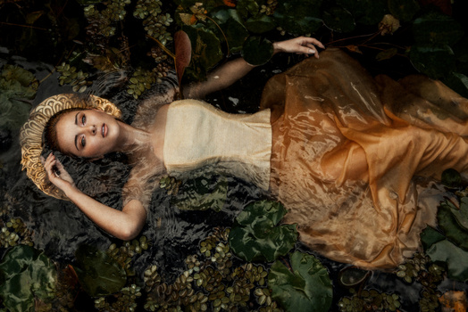 Woman in cream and gold dress lying on moss-covered ground surrounded by lush foliage.