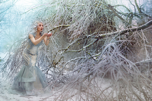 Woman in flowing gray dress posing among frost-covered tree branches in winter.